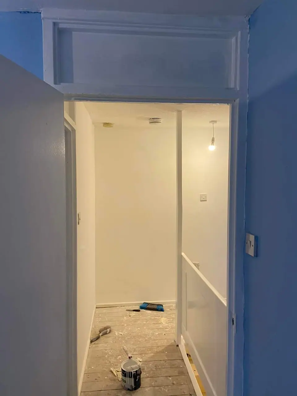 A hallway doorway with blue-painted walls and woodwork, bare floorboards and a paint tin